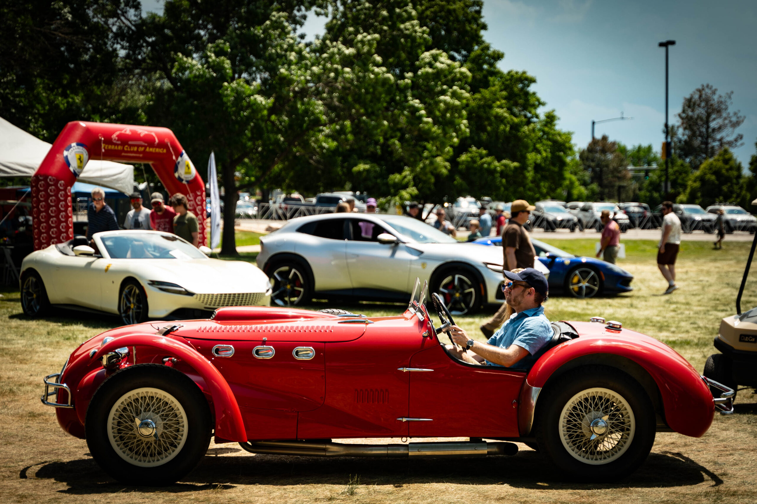 Rare classic car passes in front of Ferraris and festival attendees on a warm day in June.